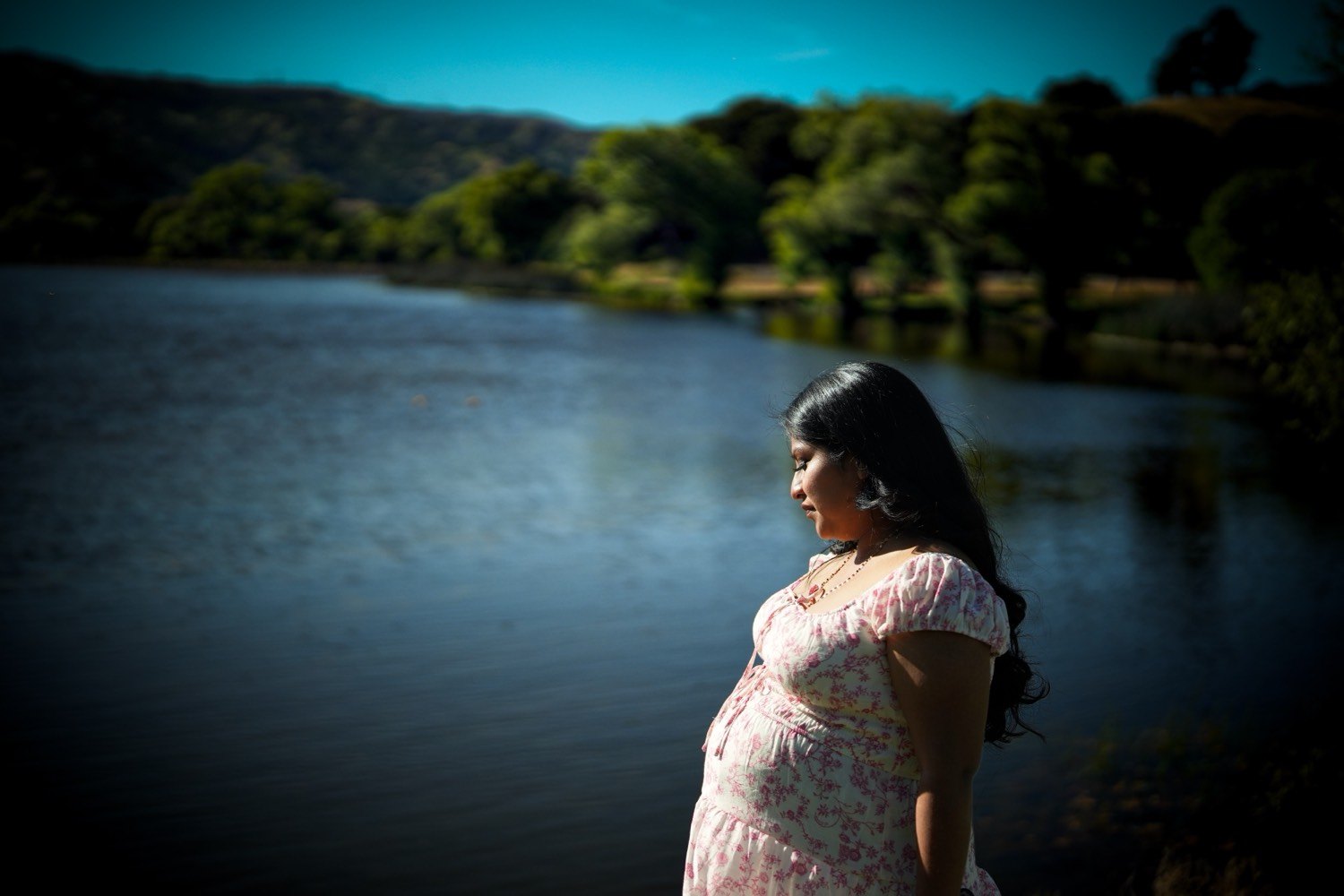 The same woman in profile at the edge of a lake, hills and trees behind her