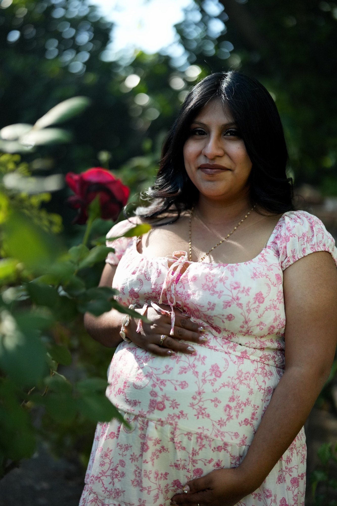A pregnant woman in a white dress with pink flowers, one hand on her belly, standing beside a red rose