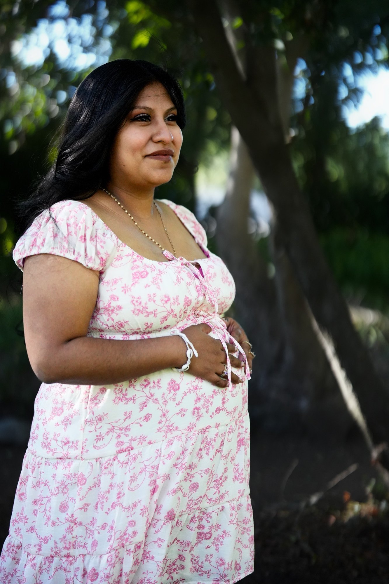 The same woman in the pink floral dress, looking aside in the trees with both hands resting on her belly
