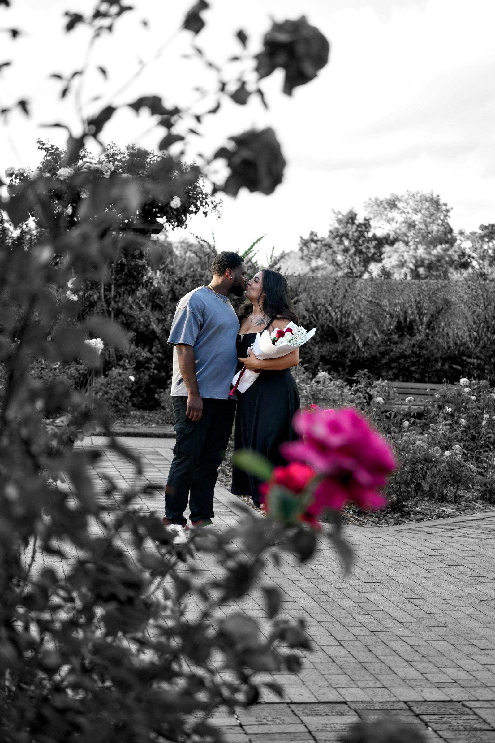 A couple kissing on a garden path, the woman holding a rose bouquet, with a pink rose in the foreground