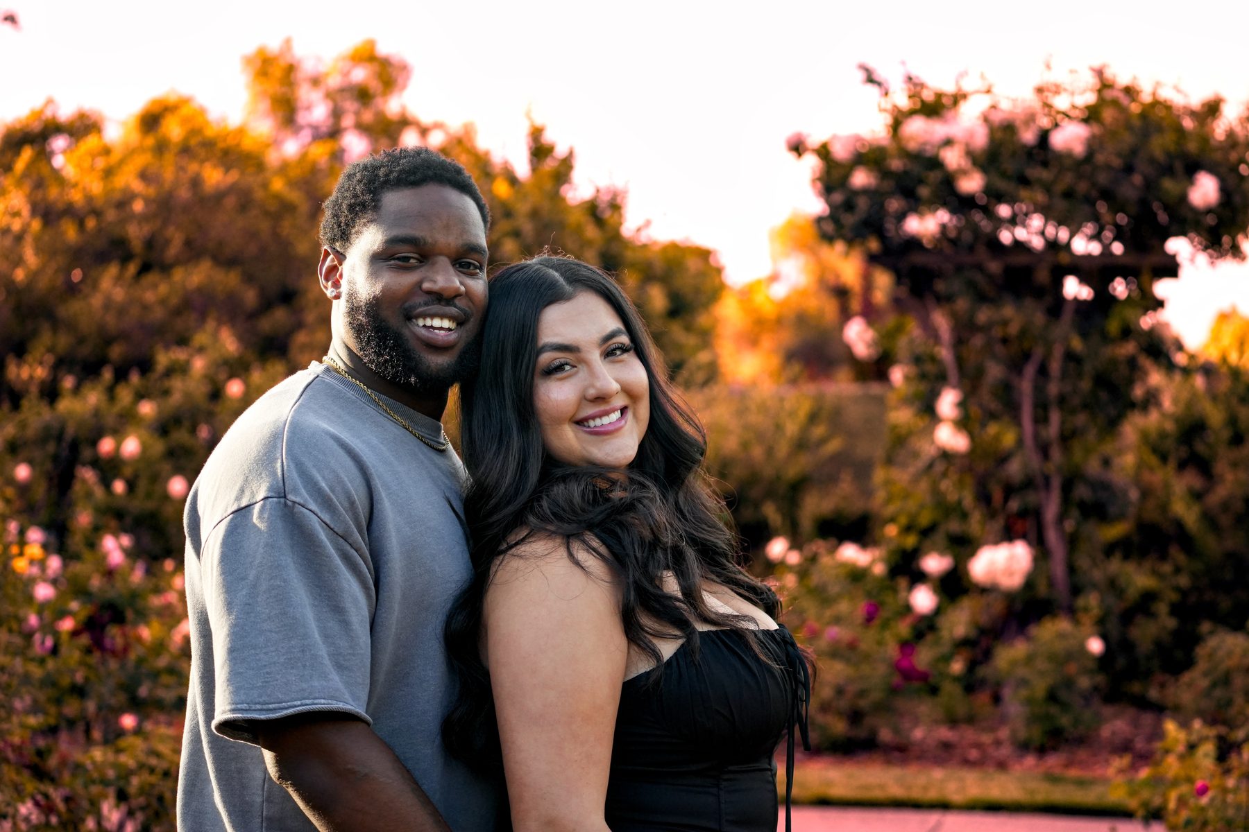 A couple smiling in a rose garden at sunset, her head resting against his shoulder