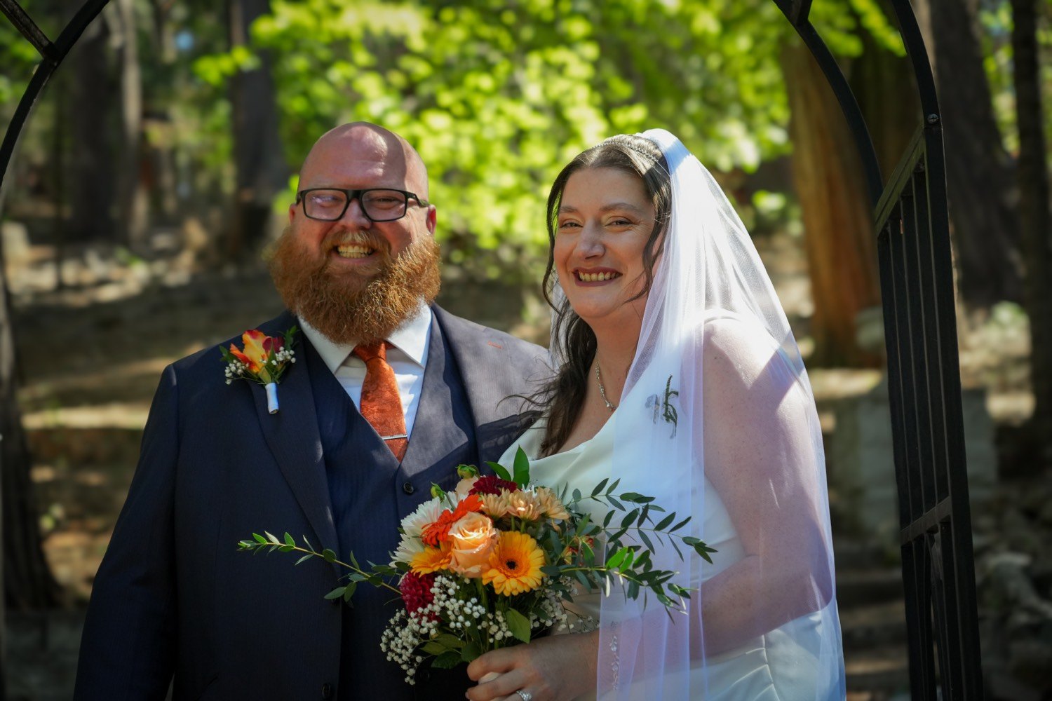 A bride in a white gown and veil and a groom with a red beard, glasses, navy vest, and rust tie, smiling under a black garden arch