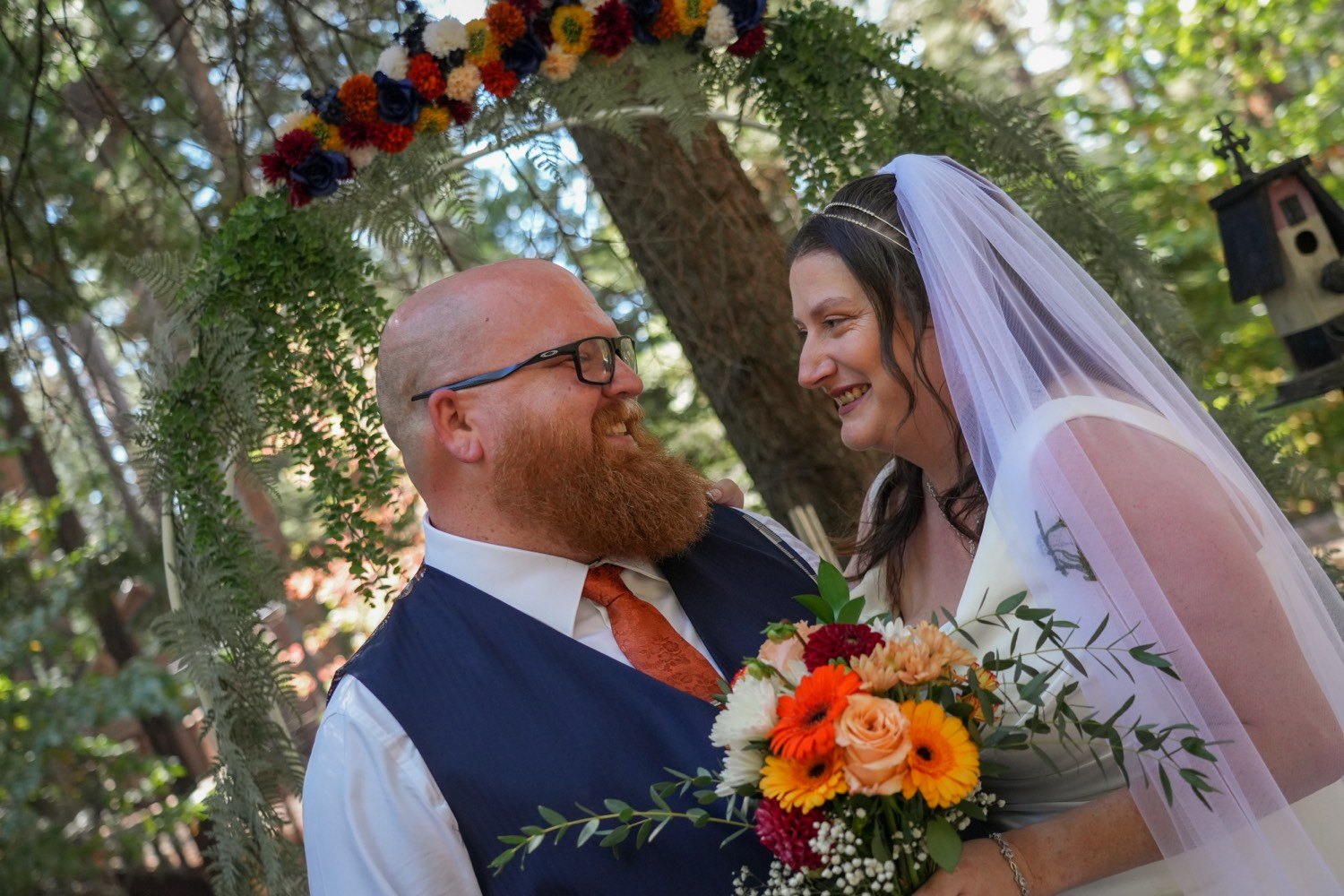 The same couple looking at each other under a floral arch of orange, red, and blue flowers in the woods